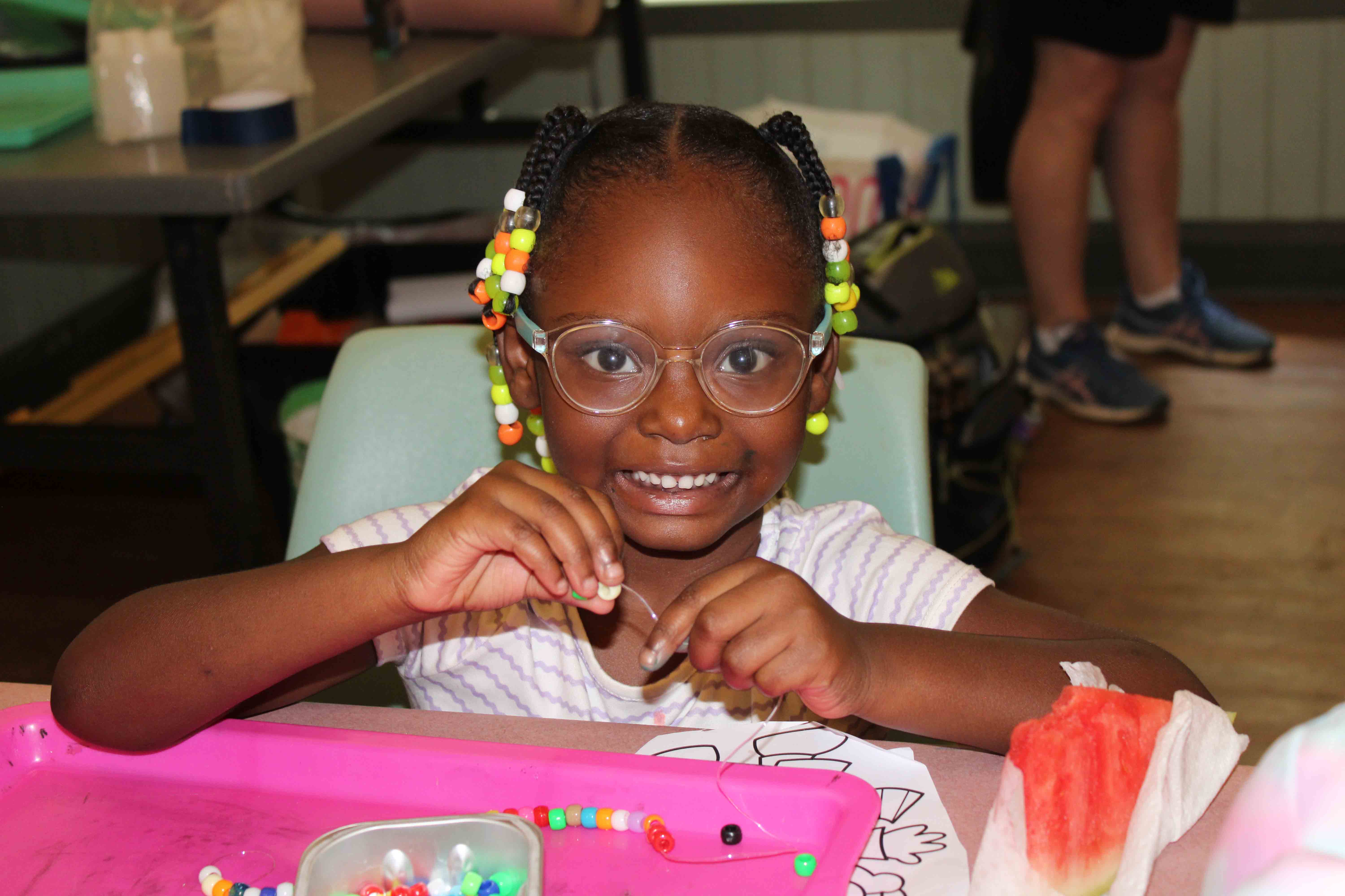 A smiling young camper with braided hair and colorful beads making a custom bracelet next to a slice of fresh watermelon.